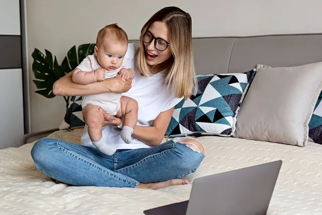 Frau mit Baby auf dem Bett schaut gemeinsam auf Laptop, Familie und Babyzeit.
