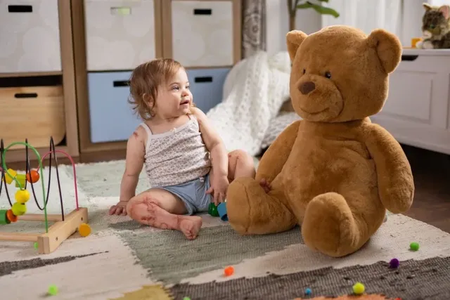 Kleines Kind sitzt mit großem Teddybär auf dem Boden im Spielzimmer mit buntem Holzspielzeug.