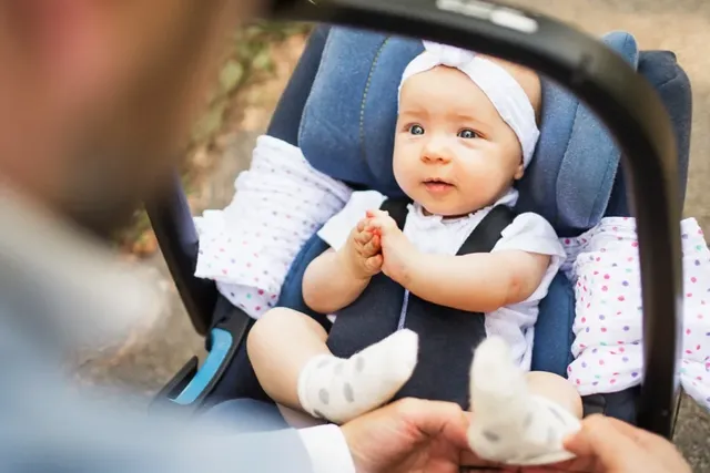 Baby im Autositz lächelt und wird von Elternteil gehalten.