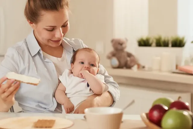 Mutter hält Baby im Arm und frühstückt gemeinsam in der Küche.