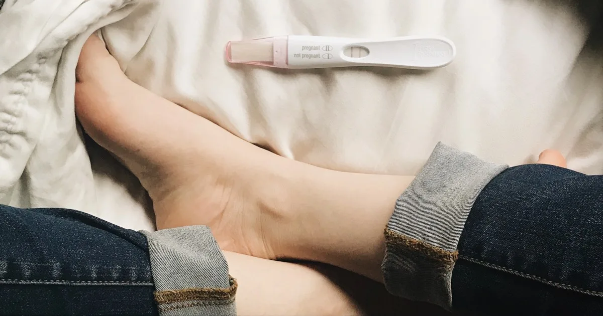 Positiver Schwangerschaftstest liegt neben entspannten Füßen auf hellem Bett.