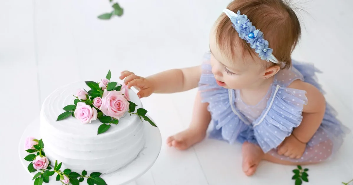 Kleines Mädchen in blauem Kleid berührt neugierig eine weiße Torte mit rosa Rosen.