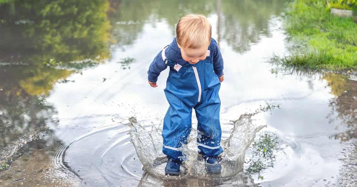 Kleinkind in blauer Regenkleidung spielt fröhlich in Wasserpfütze draußen.