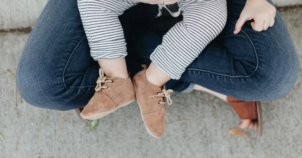Babyfüße mit braunen Schuhen auf dem Schoß eines Erwachsenen in Jeans.