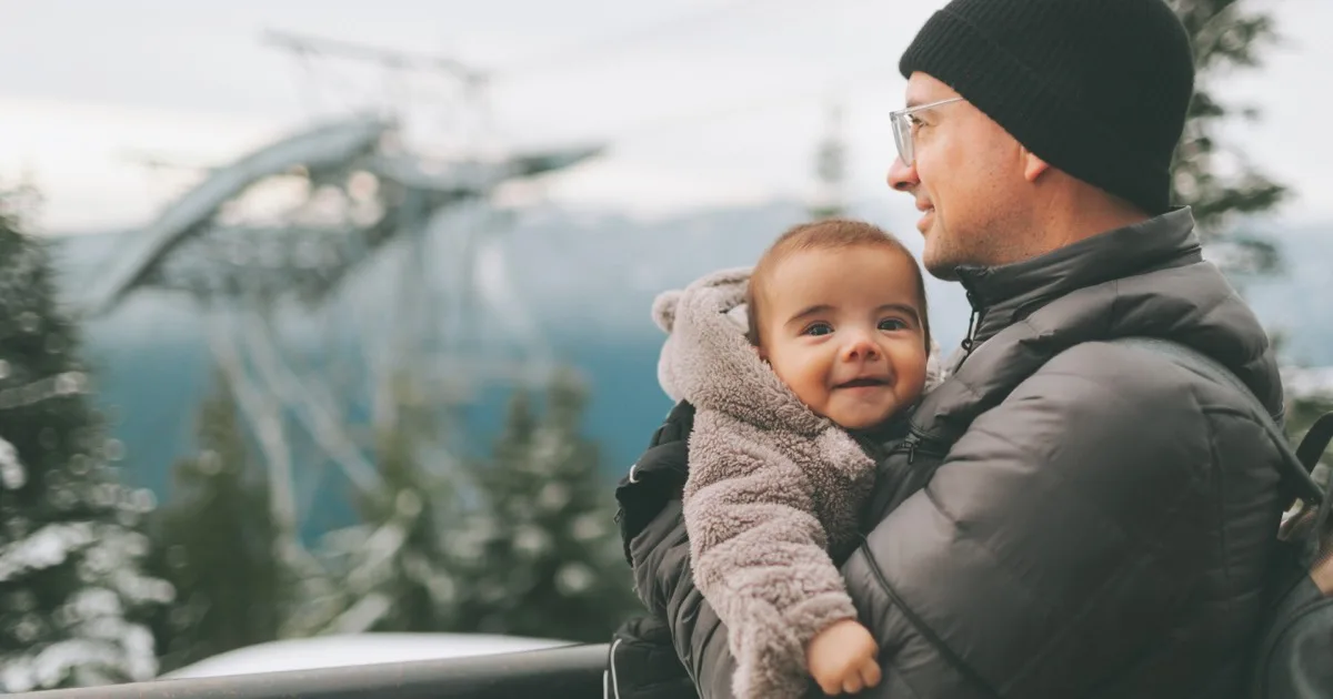 Vater hält lächelnden Baby in Winterkleidung im Arm, draußen in der Natur.