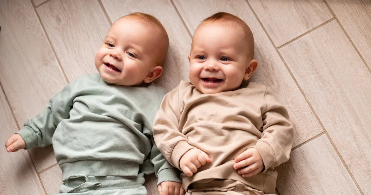 Zwei lächelnde Babys liegen auf dem Boden in bequemer Kleidung.