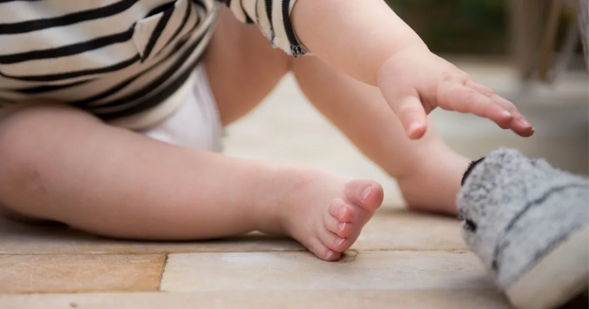 Baby sitzt auf dem Boden und greift nach einem grauen Schuh.