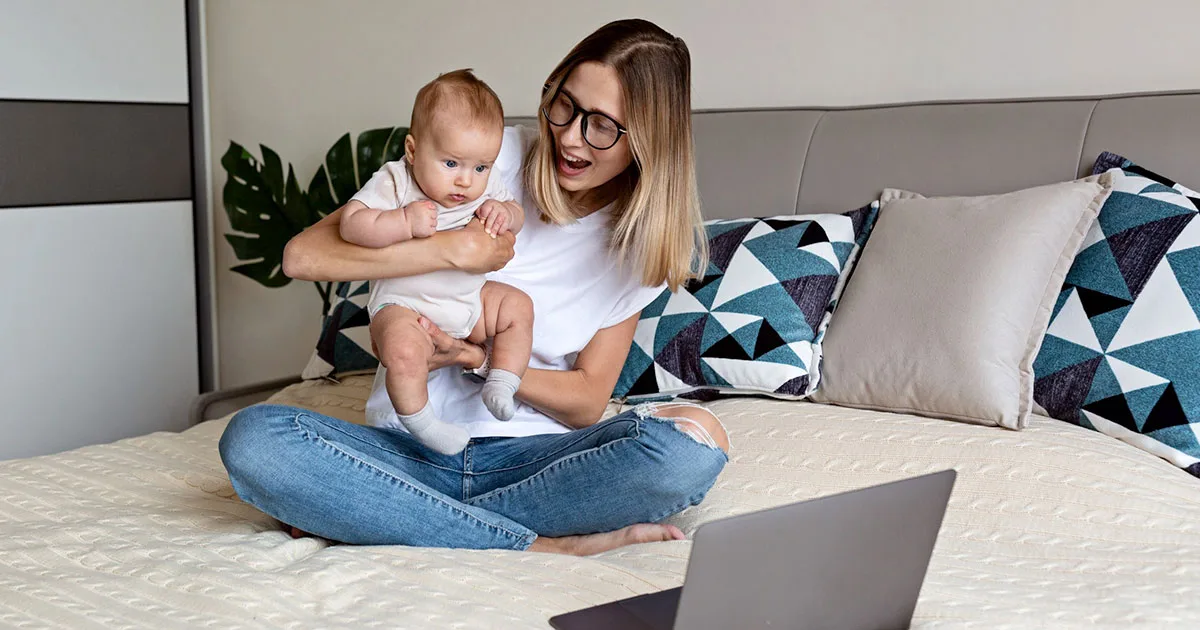 Frau mit Baby auf dem Bett schaut gemeinsam auf Laptop, Familie und Babyzeit.