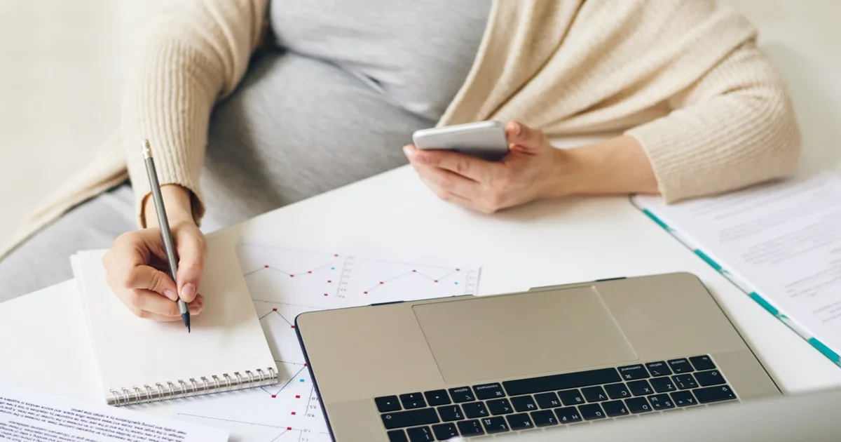 Schwangere Frau arbeitet am Laptop und macht Notizen mit Smartphone in der Hand.