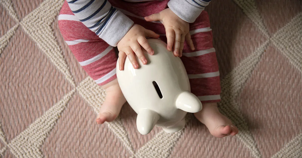 Baby sitzt auf Teppich und hält ein weißes Sparschwein.