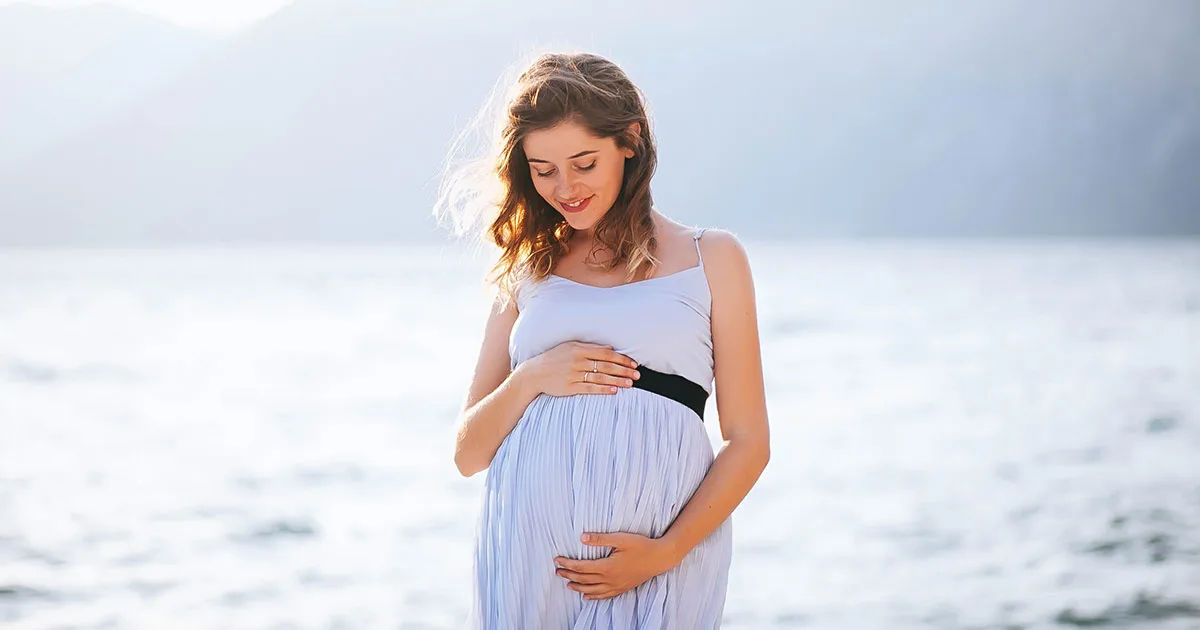 Schwangere Frau am Wasser mit Händen auf dem Babybauch, glücklich und entspannt.