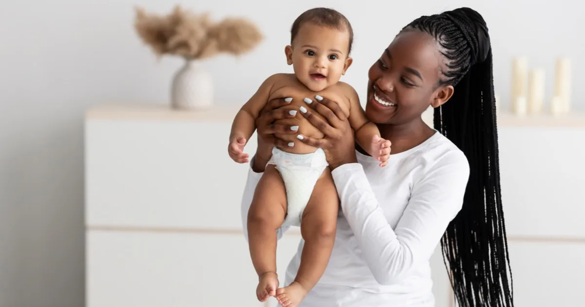 Mutter hält lächelnd ihr Baby in den Armen in einem hellen Wohnzimmer.