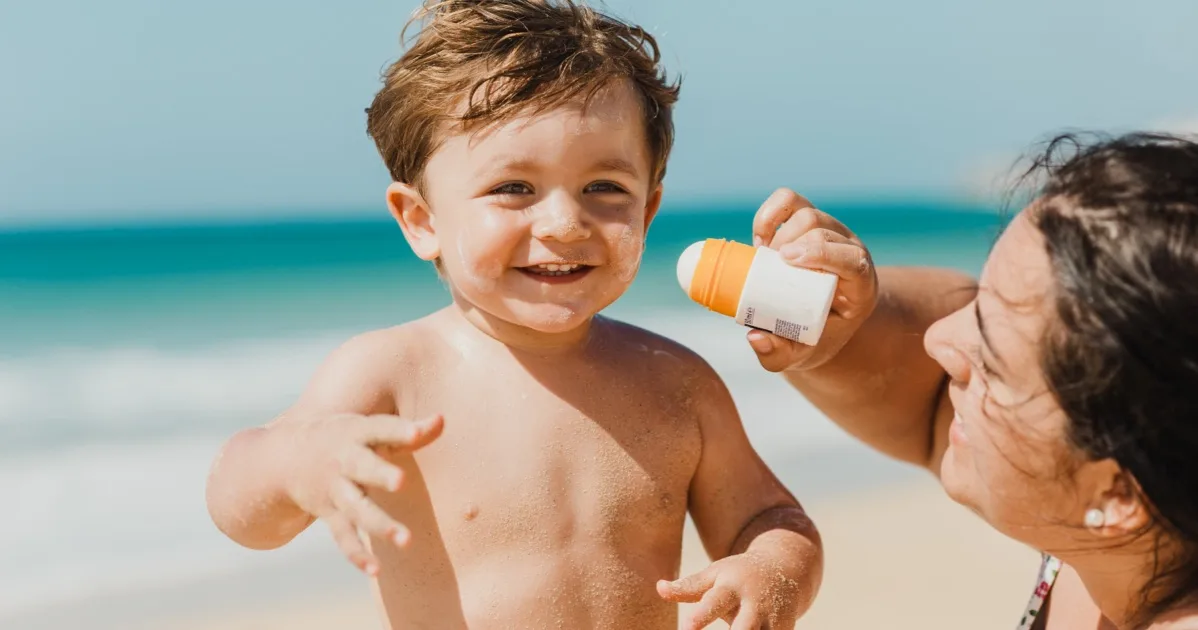 Kind am Strand bekommt Sonnencreme von Mutter aufgetragen, fröhliche Familienszene